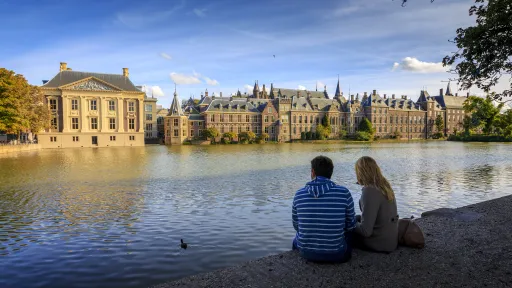 Two people sitting by the Hofvijver in The Hague with the Marutishuis and the Binnenhof in the background.