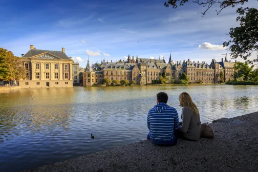 Two people sitting by the Hofvijver in The Hague with the Marutishuis and the Binnenhof in the background.