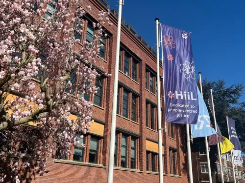 A HiiL vertical banner is flapping on a pole in front of a brown building with lots of windows, it reads "dedicated to people-centered justice". On the left, a tree in full bloom