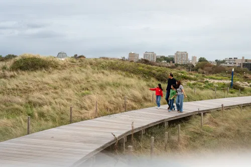 A family walking on a wooden path between grassy dunes. In the background, the city of The Hague.
